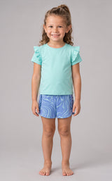 Toddler girl wearing a light turquoise short sleeve top with ruffled shoulders and blue patterned running shorts.