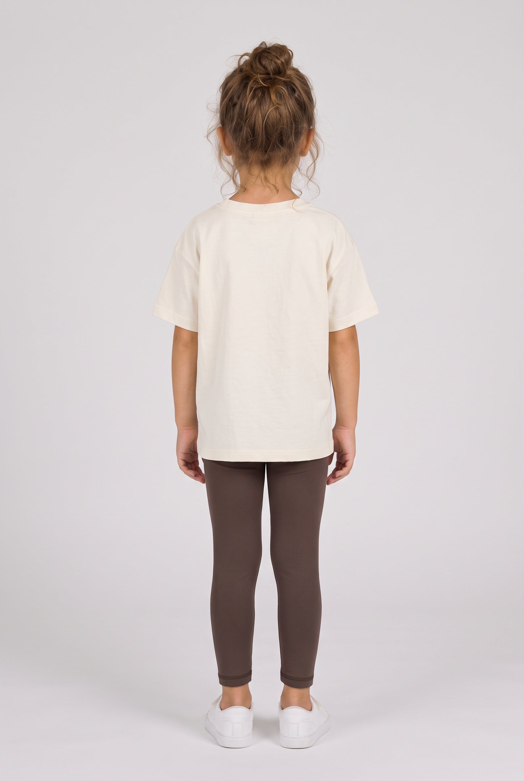 Toddler wearing slim dark gray leggings with a cream short-sleeve top and white sneakers, shown from behind.