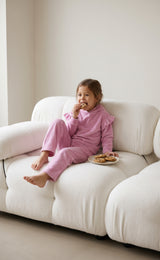 Little girl wearing a pink velour hoodie and flared pants, seated on a white sofa with a snack plate.