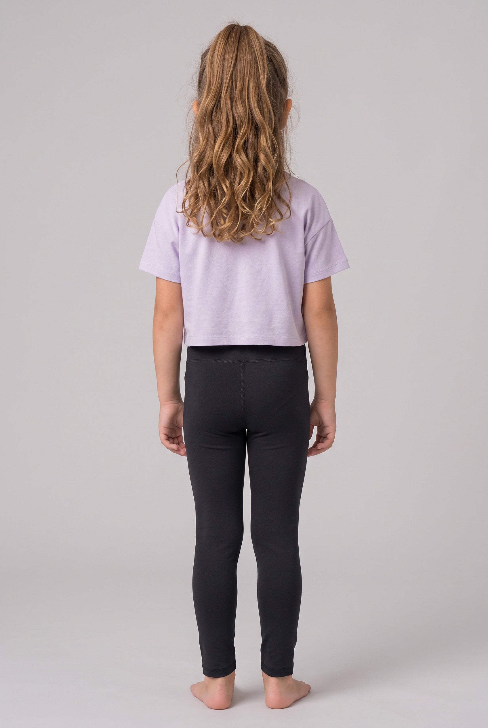 A girl models full-length black leggings with a lavender short-sleeve top, shown from behind.
