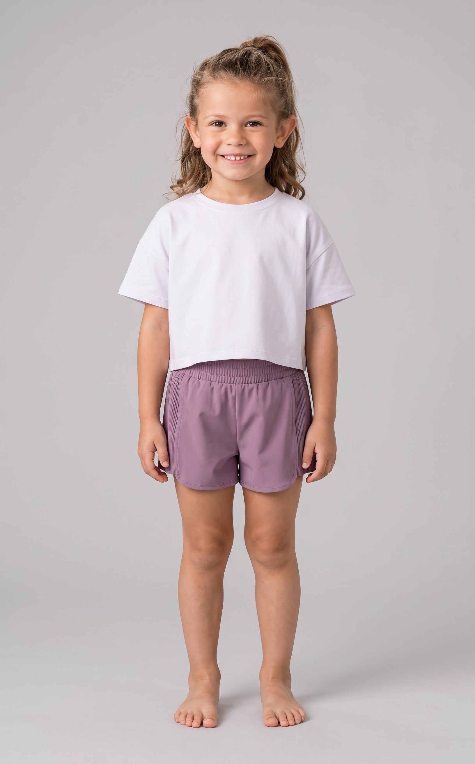 Toddler girl wearing dusky mauve lightweight shorts with elastic waistband and a plain white short-sleeve t-shirt, standing.
