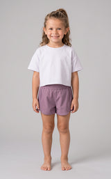 Toddler girl wearing dusky mauve lightweight shorts with elastic waistband and a plain white short-sleeve t-shirt, standing.