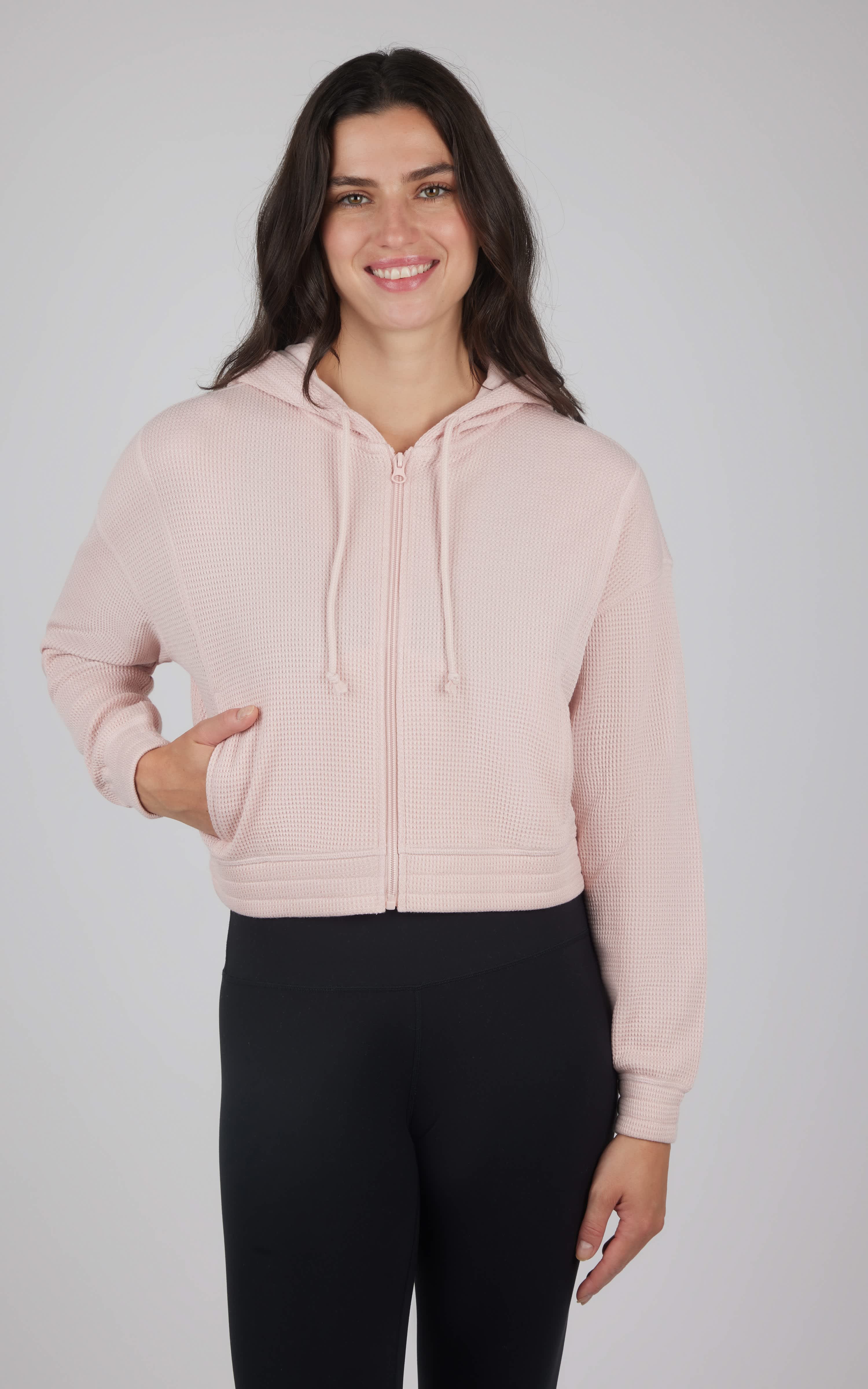 Light pink cropped hooded jacket with front pockets and drawstring hood worn with black pants.