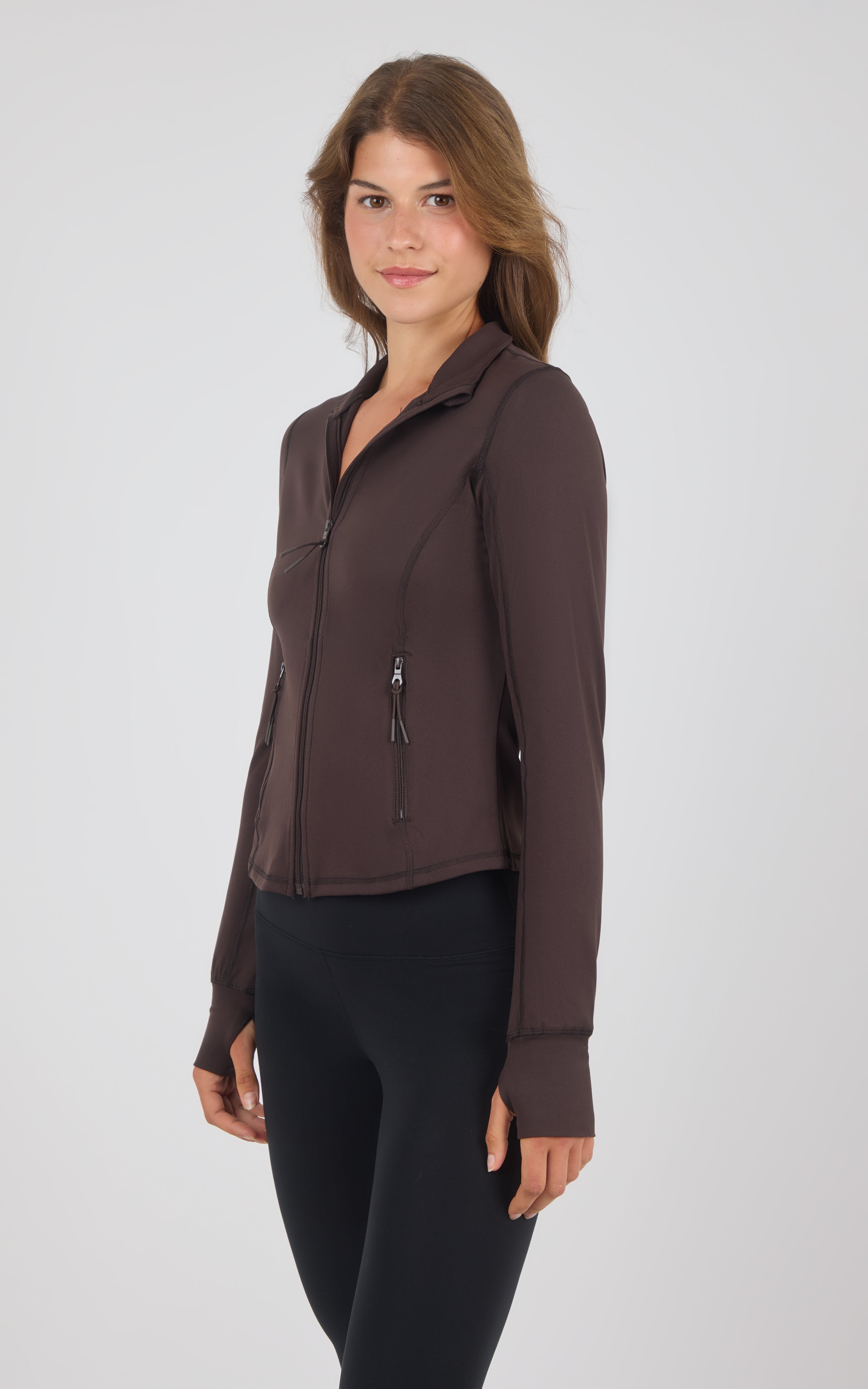A woman modeling a dark brown long-sleeve athletic zip-up jacket with a stand collar and zippered pockets, paired with black leggings, against a light gray studio background.
