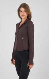 A woman modeling a dark brown long-sleeve athletic zip-up jacket with a stand collar and zippered pockets, paired with black leggings, against a light gray studio background.