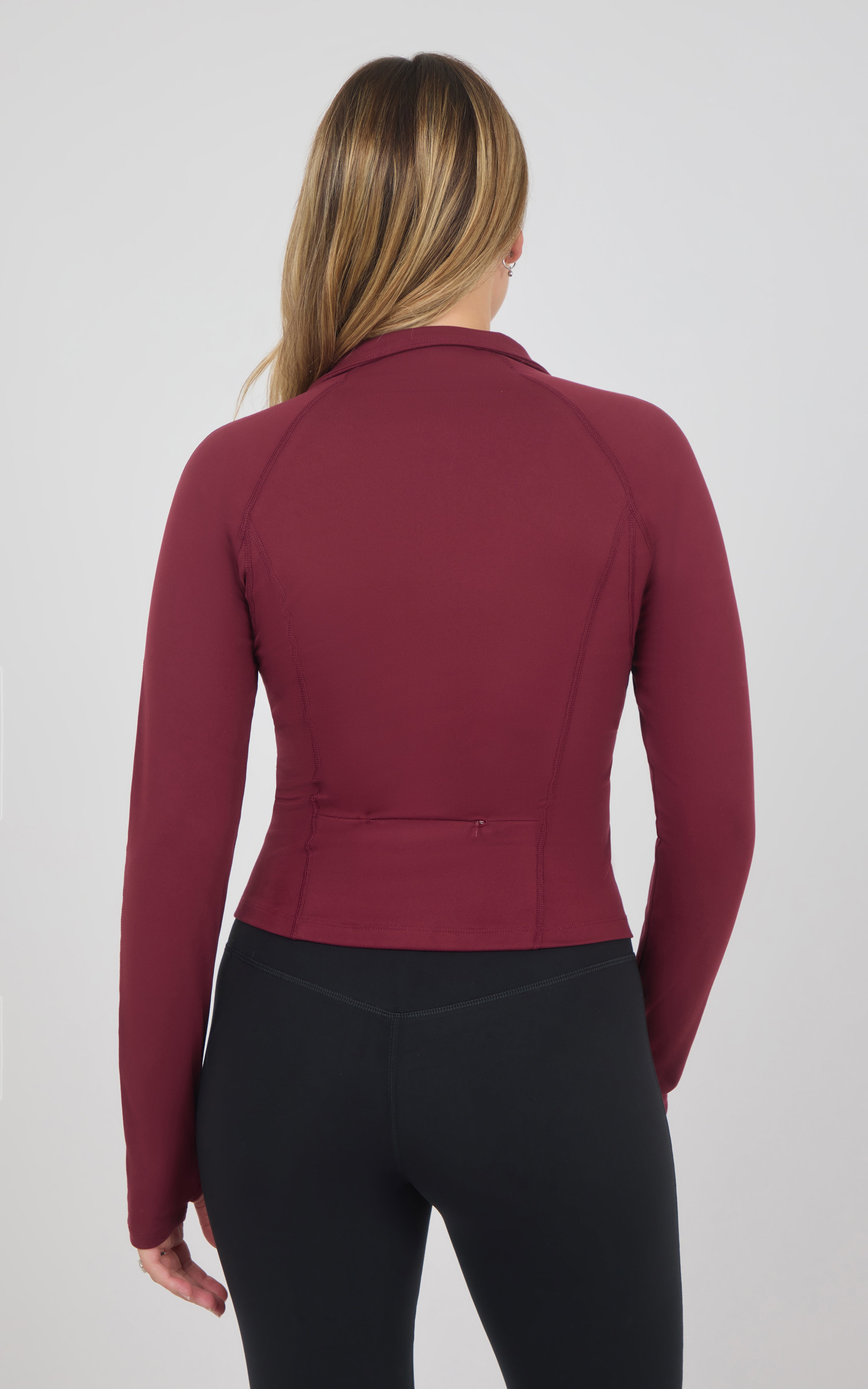 Back view of a burgundy long-sleeve raglan jacket with a raised collar and fitted waist, paired with black pants.