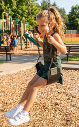 Girl wearing a dark green sleeveless top and matching crossover shorts, seated on a playground swing with white sneakers.