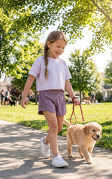 Girl in a lavender T-shirt and mauve athletic shorts walks a small golden dog on a sunny park path.
