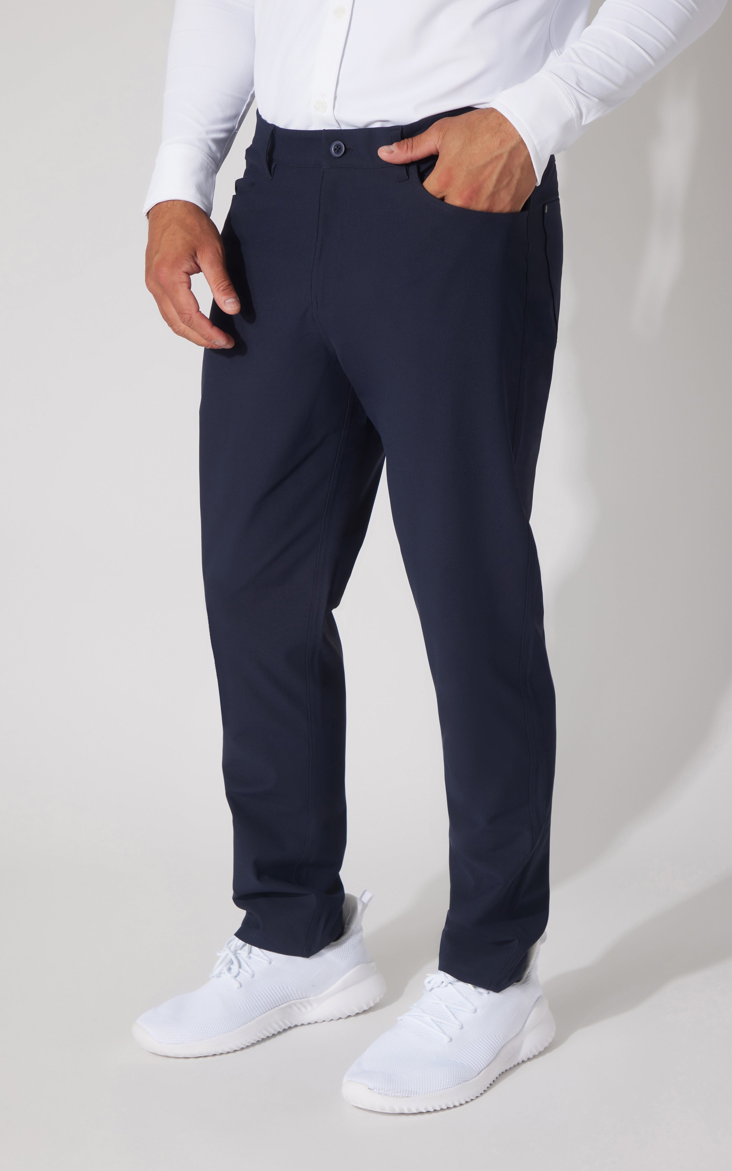 A man wearing navy blue slim-fit trousers, a white long-sleeve shirt, and white sneakers, shown from the waist down against a light gray studio background.
