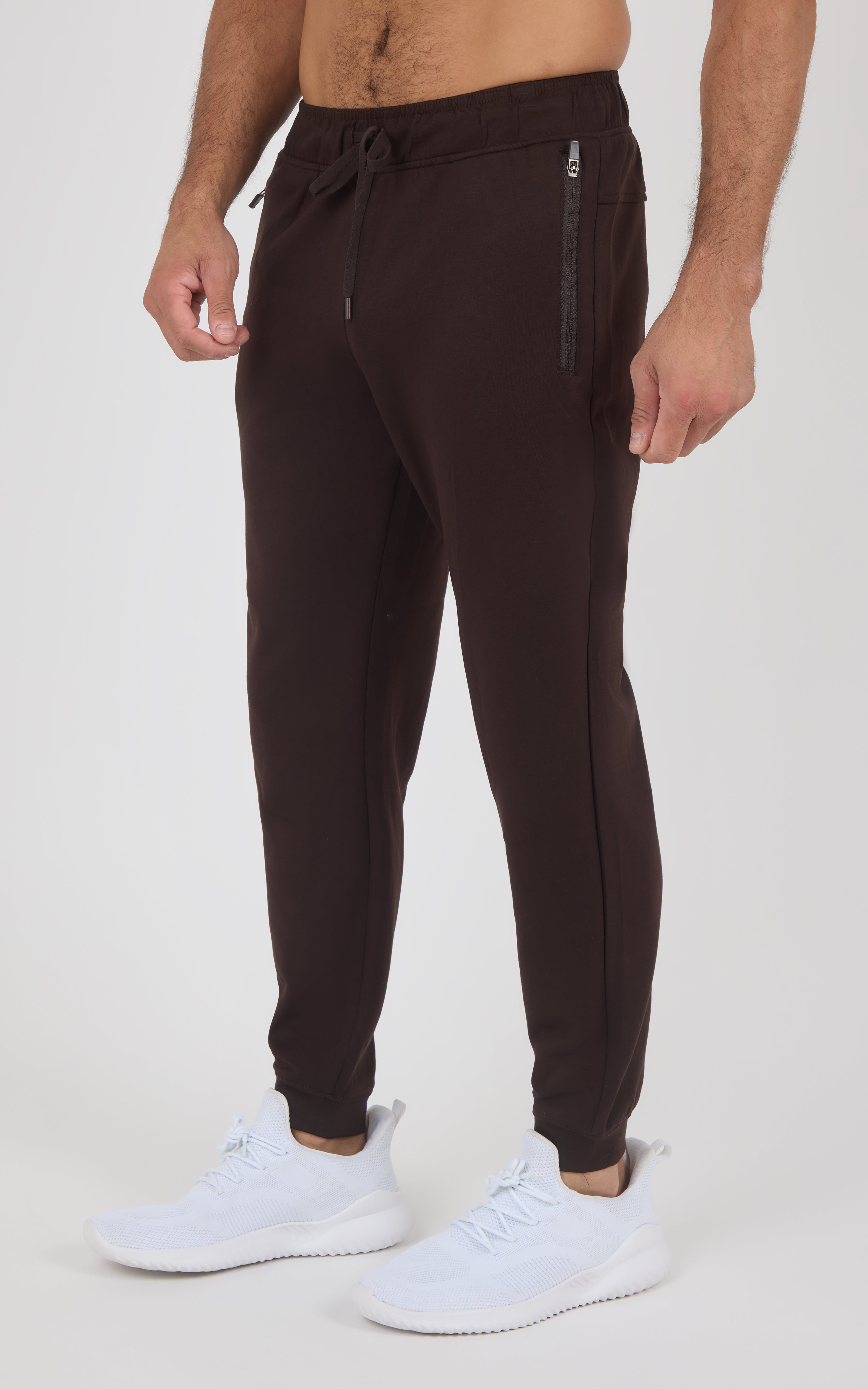 Dark brown tapered joggers with an elastic drawstring waist, zippered side pockets, and ribbed ankle cuffs, worn with white.