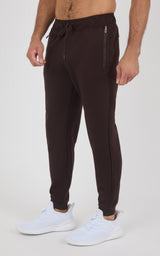Dark brown tapered joggers with an elastic drawstring waist, zippered side pockets, and ribbed ankle cuffs, worn with white.