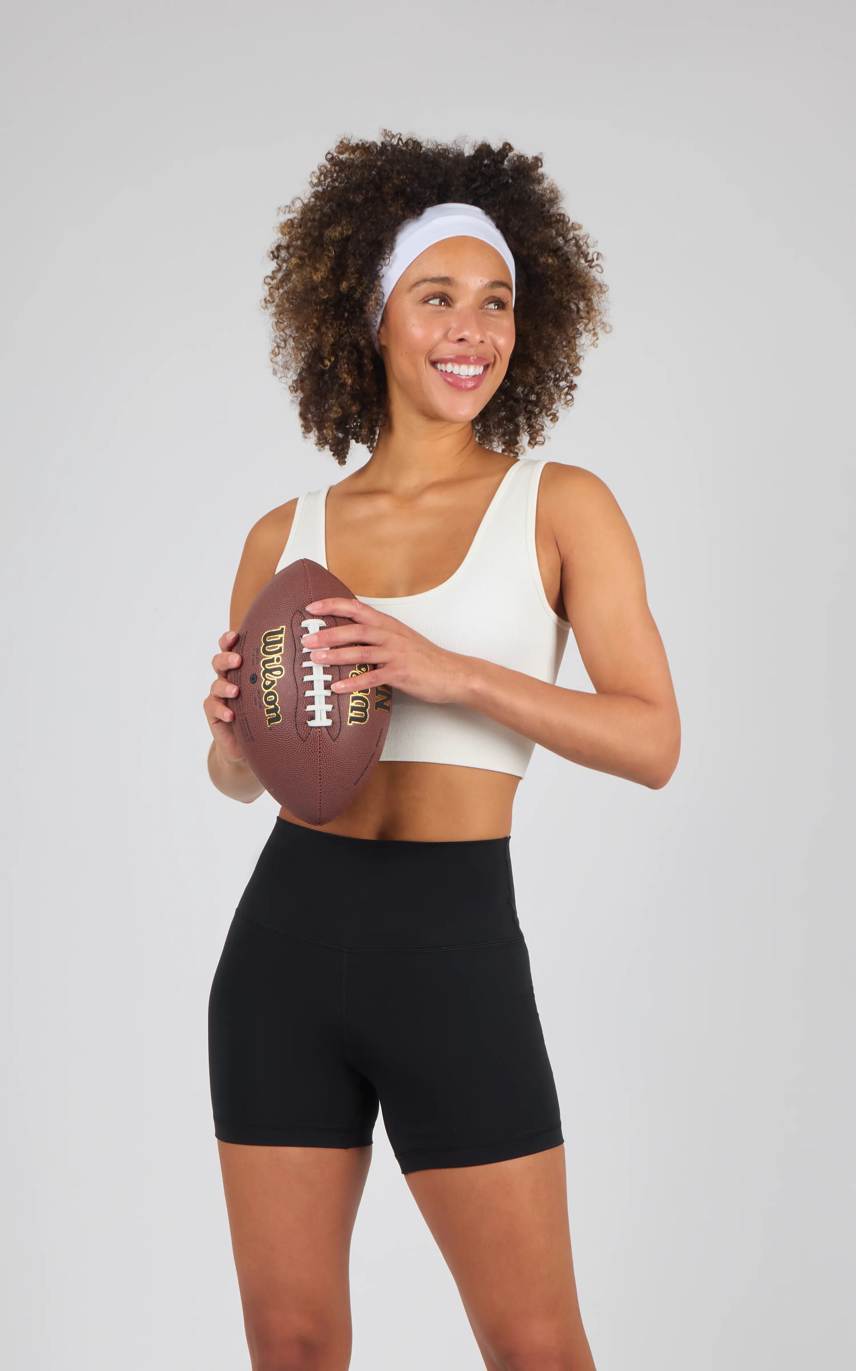 Woman wearing black 5-inch bike shorts with a high waistband and a beige crop top, holding a football and smiling.