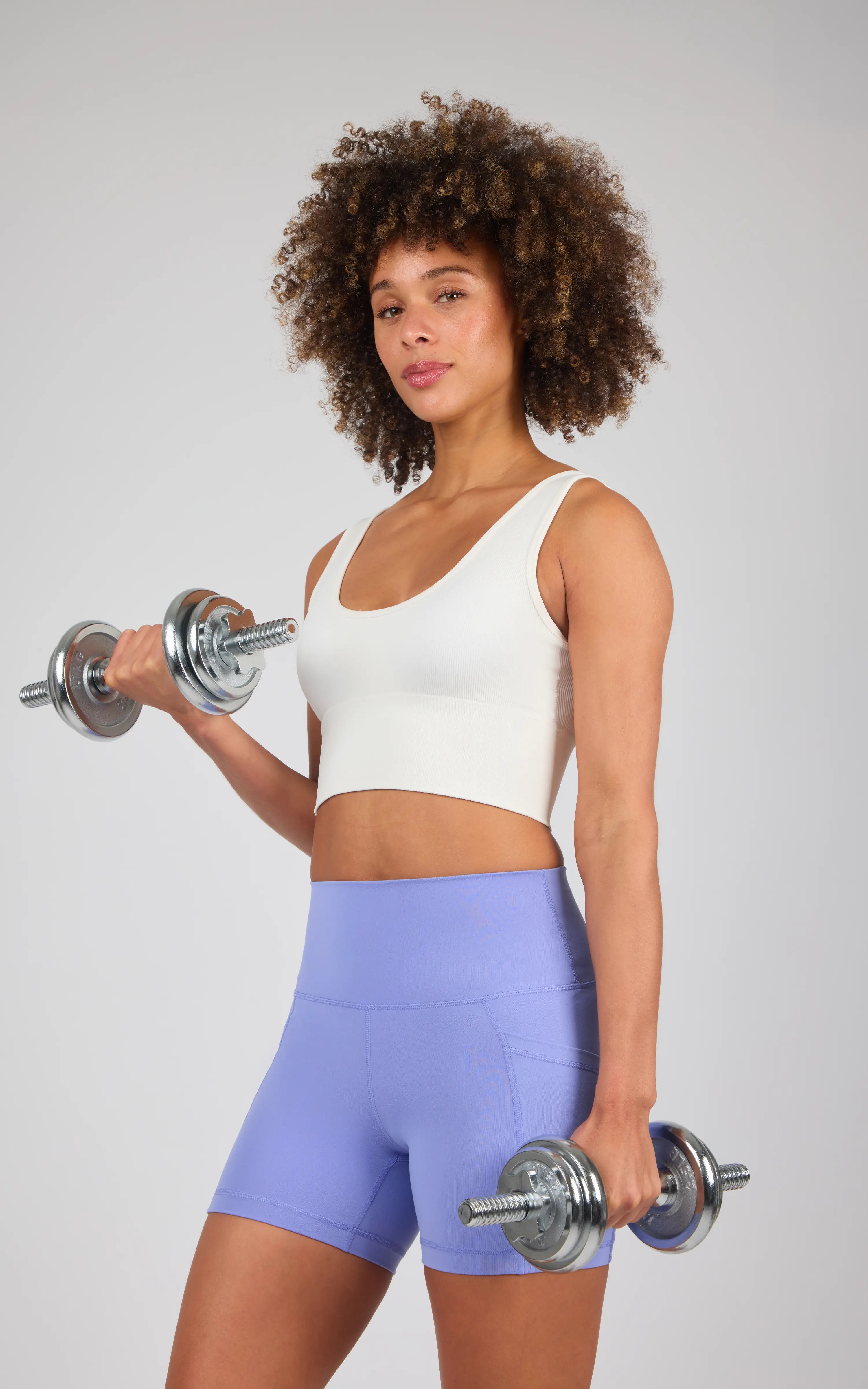 Woman wearing high-waisted, light purple 5-inch biker shorts and a white sleeveless crop top holding silver dumbbells.