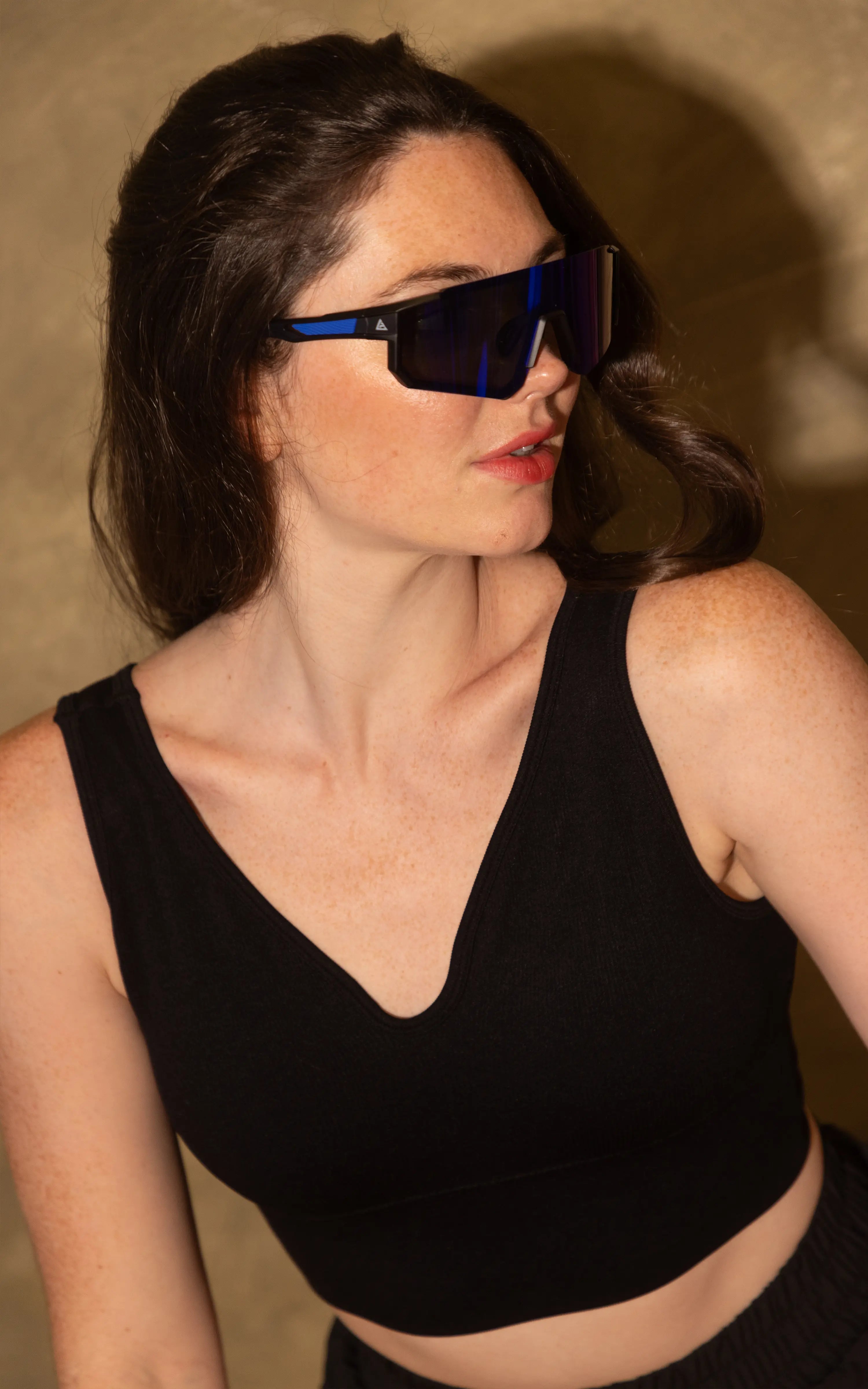 A woman wearing angular black sunglasses with bright blue temple arms and a black ribbed crop top, photographed against a warm beige textured background.