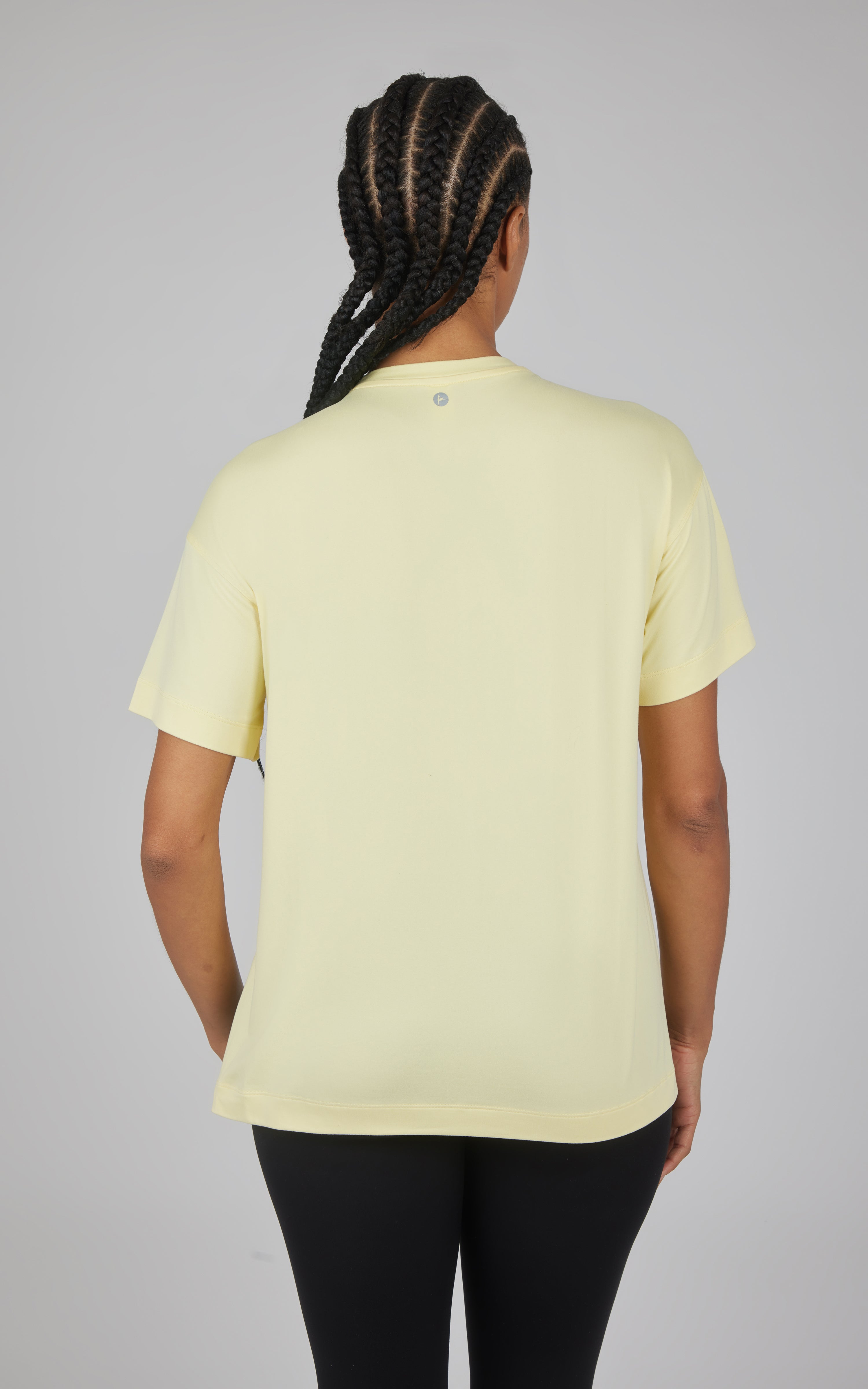 Back view of a pale yellow short-sleeve boxy top with a curved high-low hem, paired with black pants.