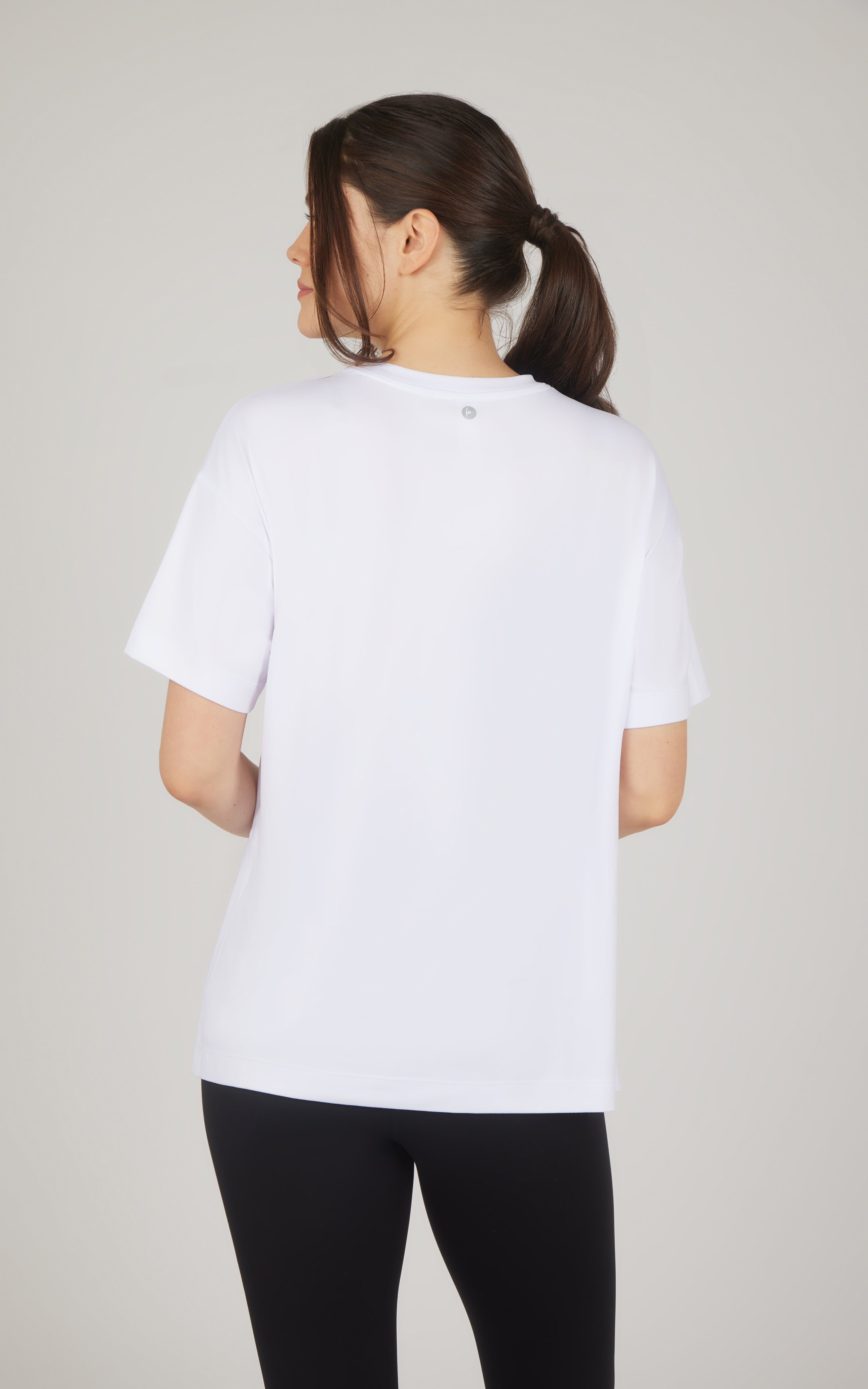 Back view of a white boxy short-sleeve top with a relaxed fit and slightly longer hem, paired with black leggings.