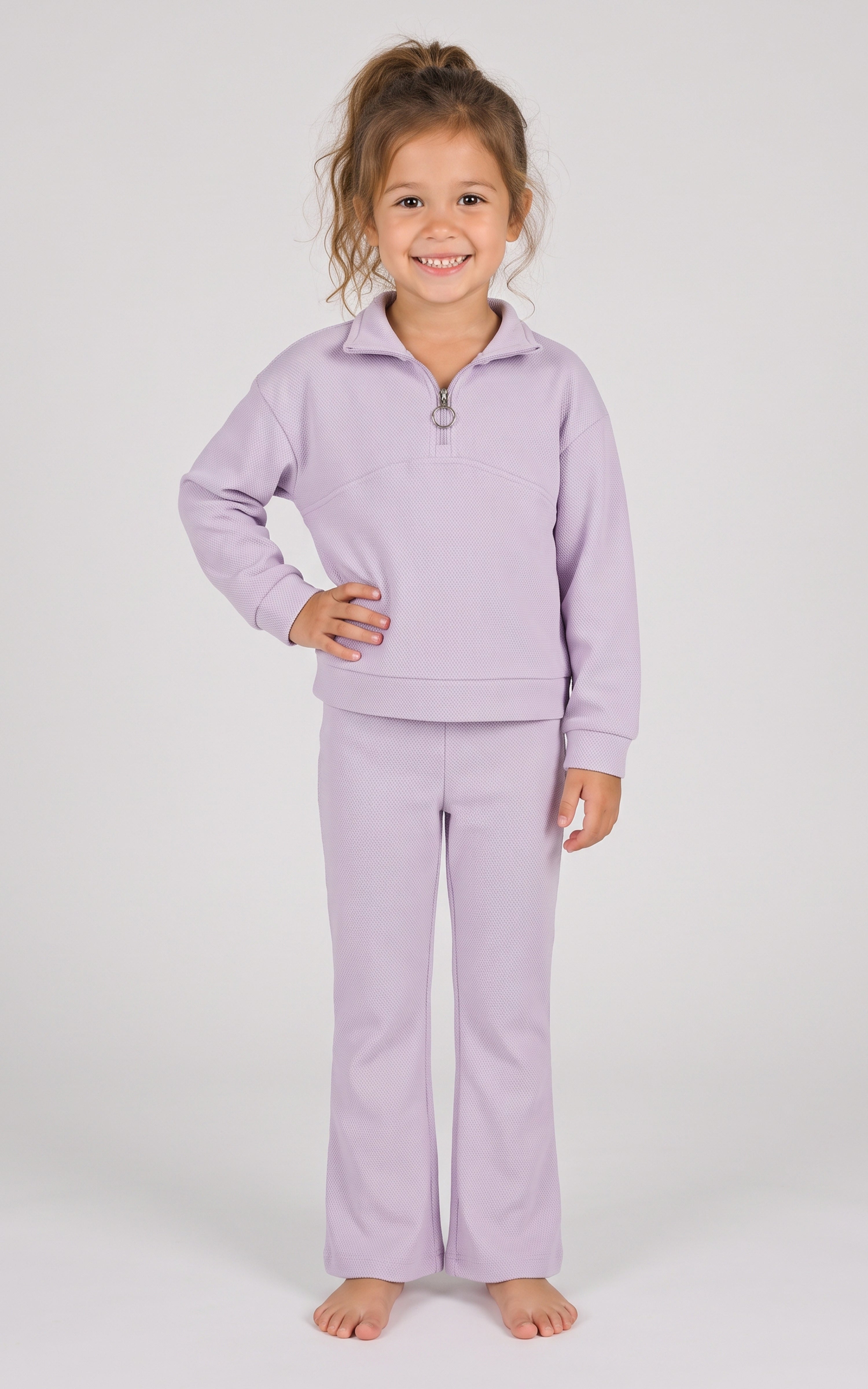 Toddler girl wearing a lavender half-zip sweatshirt and matching flared pants, standing barefoot.