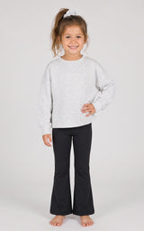 Toddler girl wearing a heather-gray long-sleeve pullover with black flared pants and a brown scrunchie in her hair.