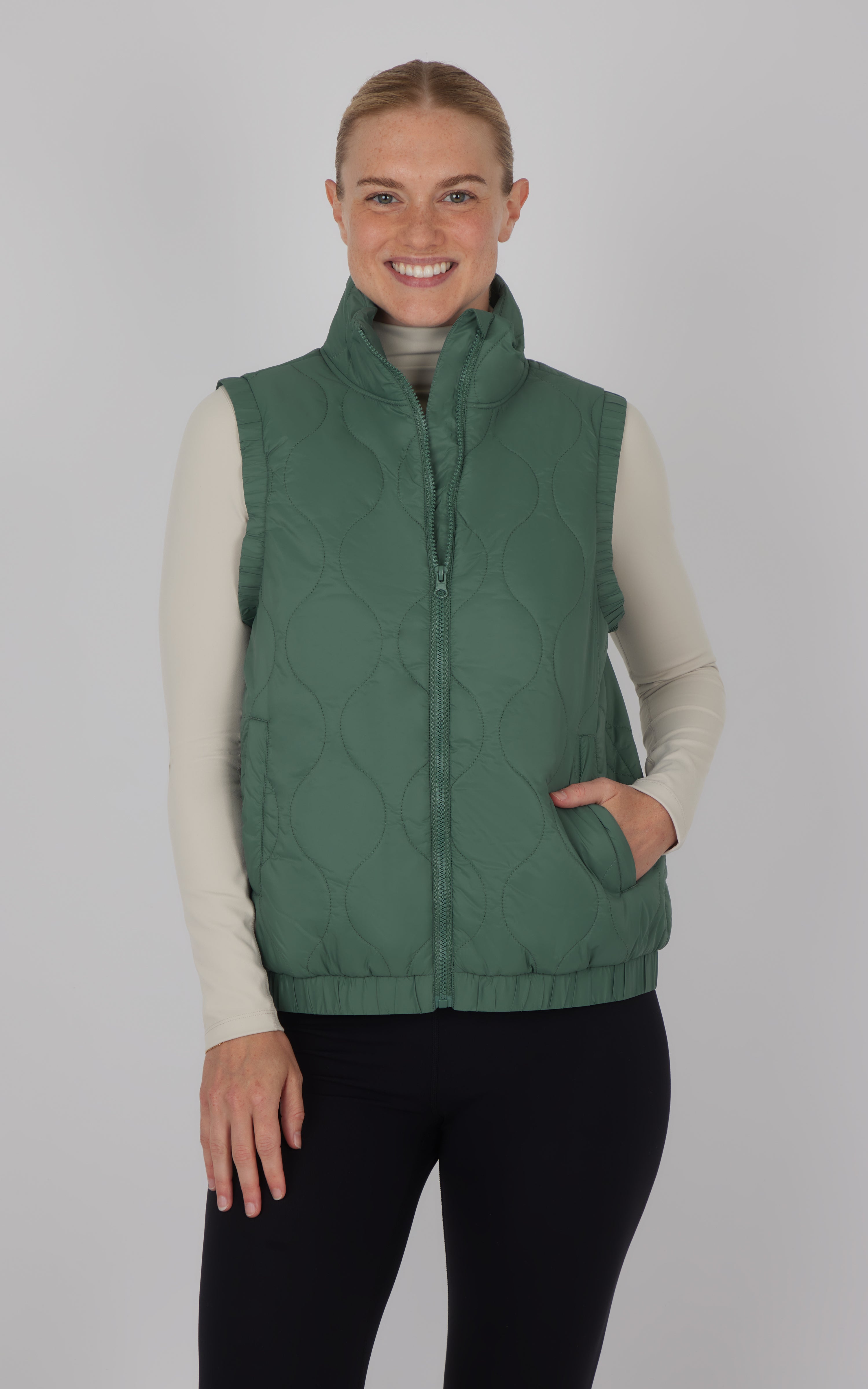 Dark forest green quilted vest with front zipper and high collar, worn over beige long-sleeve top.