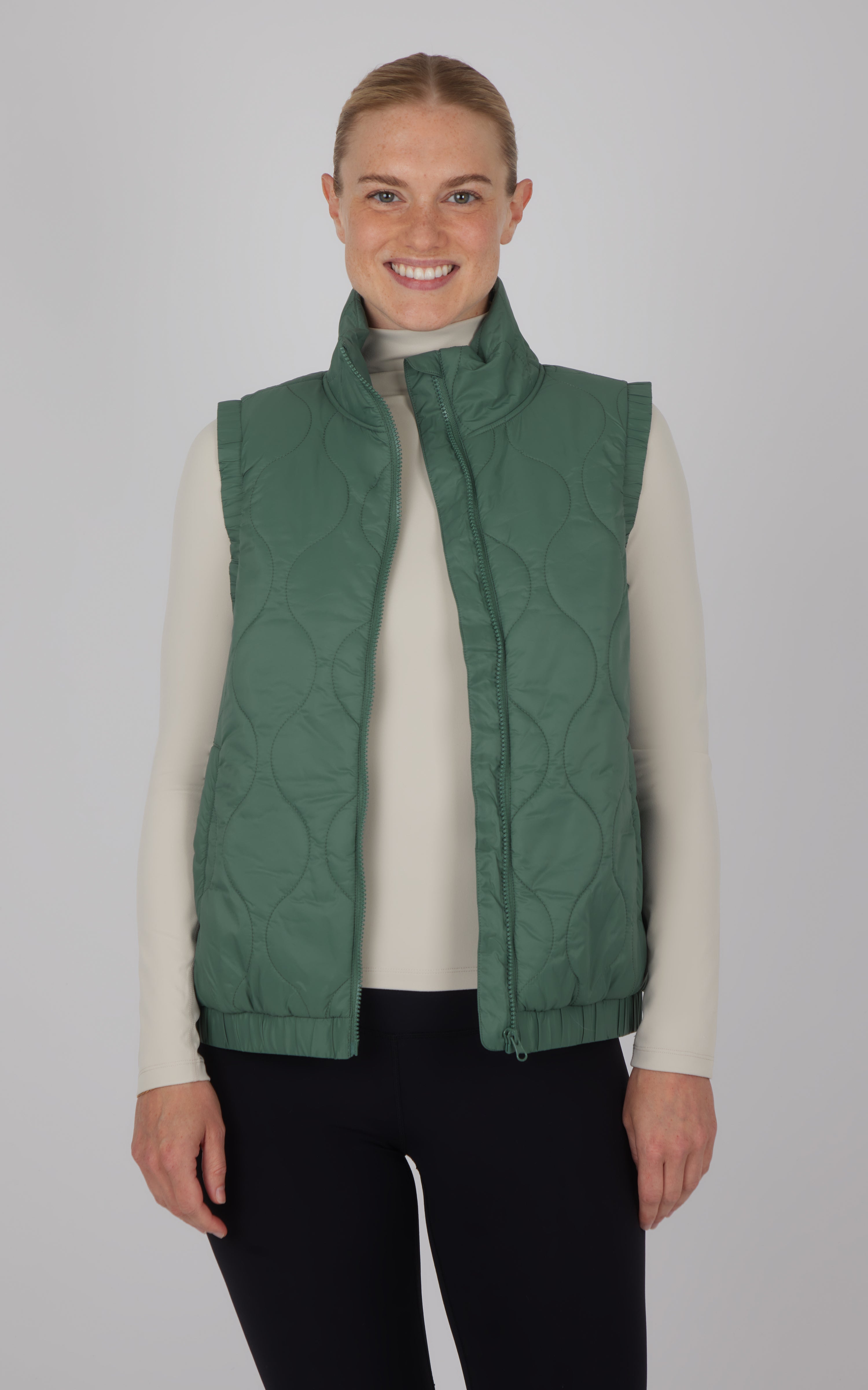 Dark forest green quilted vest with high collar, sleeveless design, worn over beige long sleeve top.