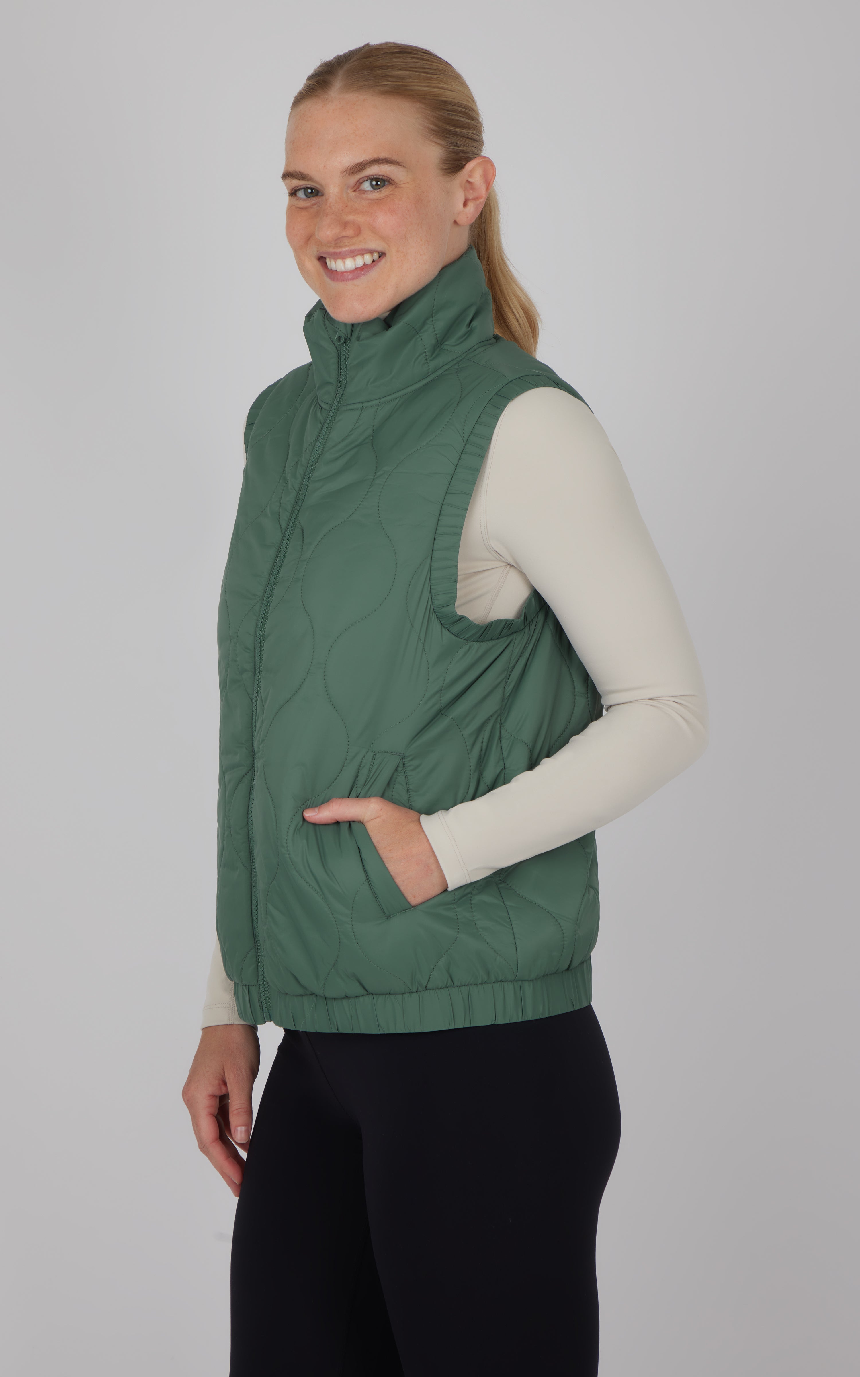 Dark forest green quilted sleeveless vest with high collar and side pockets worn over cream long sleeve top.