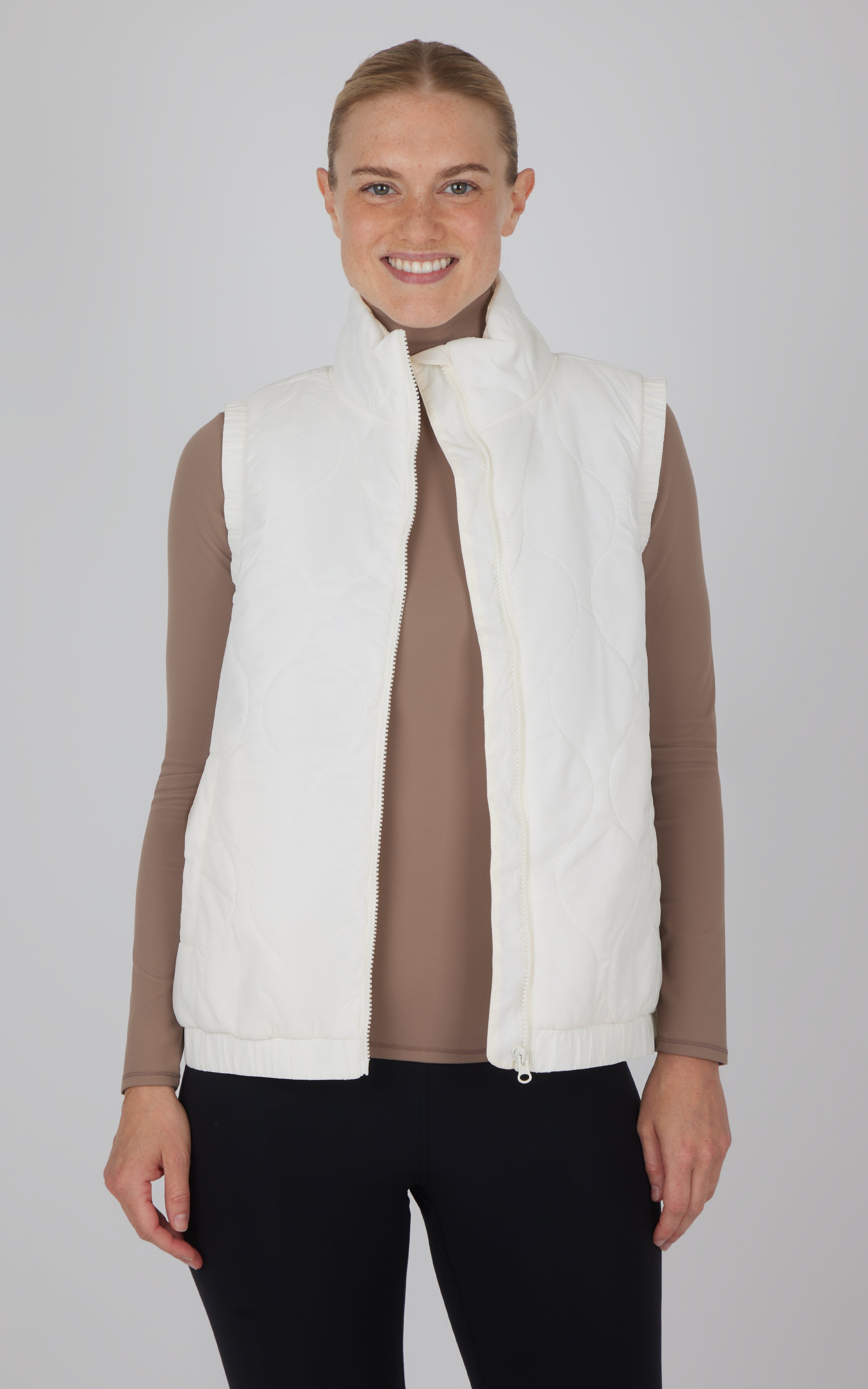 White quilted sleeveless vest with high collar, front zipper, worn over long-sleeve beige top.