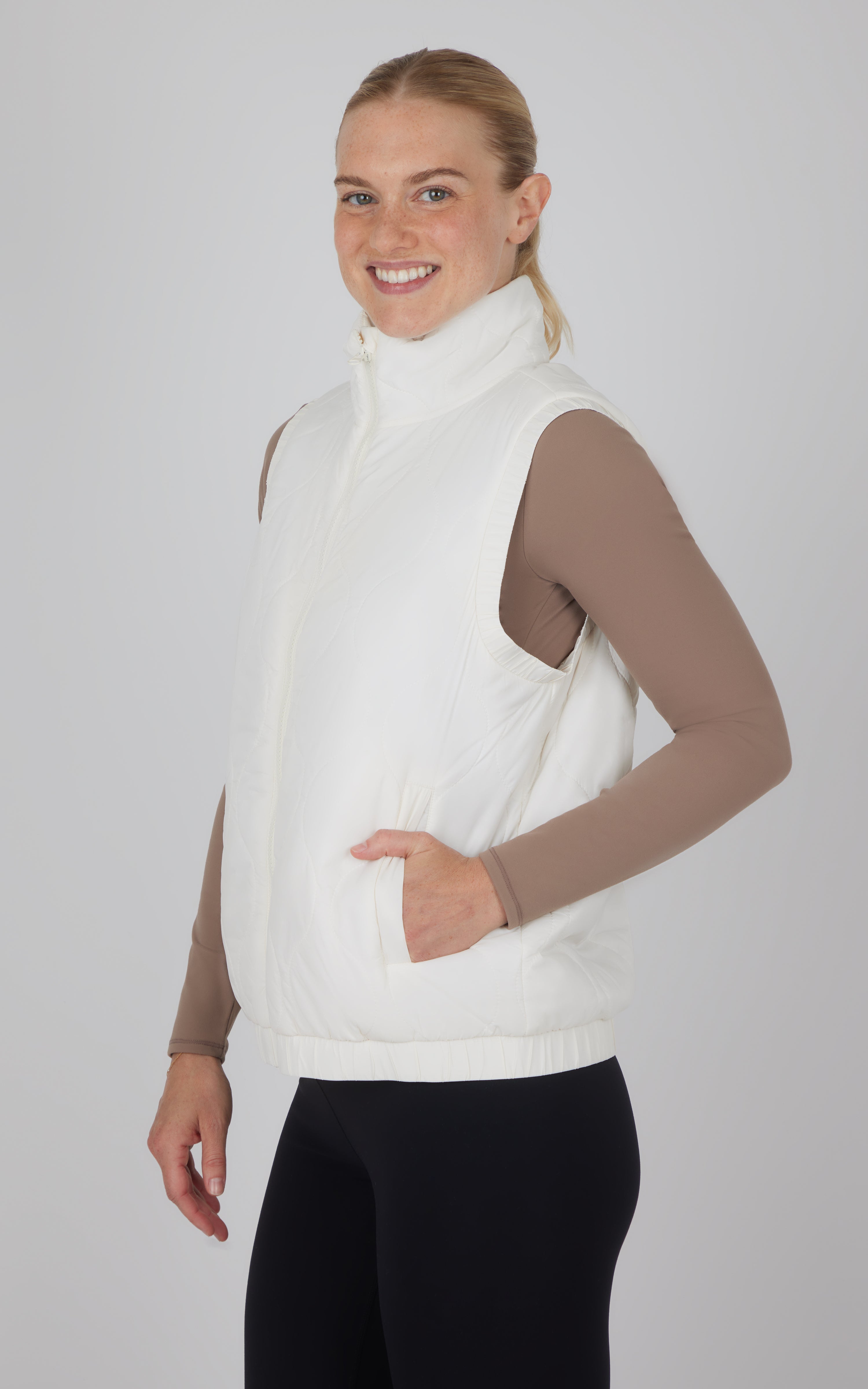 White quilted sleeveless vest with high collar and side pockets, worn over a taupe long-sleeve top.