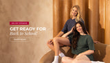 Two young women sit on a brown couch in a warm, wood-paneled room, wearing oversized T-shirts, one navy blue and one green. Promotional text reads: "25% off sitewide. Get ready for back to school. Shop now."
