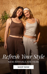 Two women posing in matching lounge sets in a warmly lit room with a textured gold wall, a curved sofa, and a potted plant. One set is dark brown and the other is light gray. Promotional text reads: "Refresh your style. new arrivals are here. shop now"