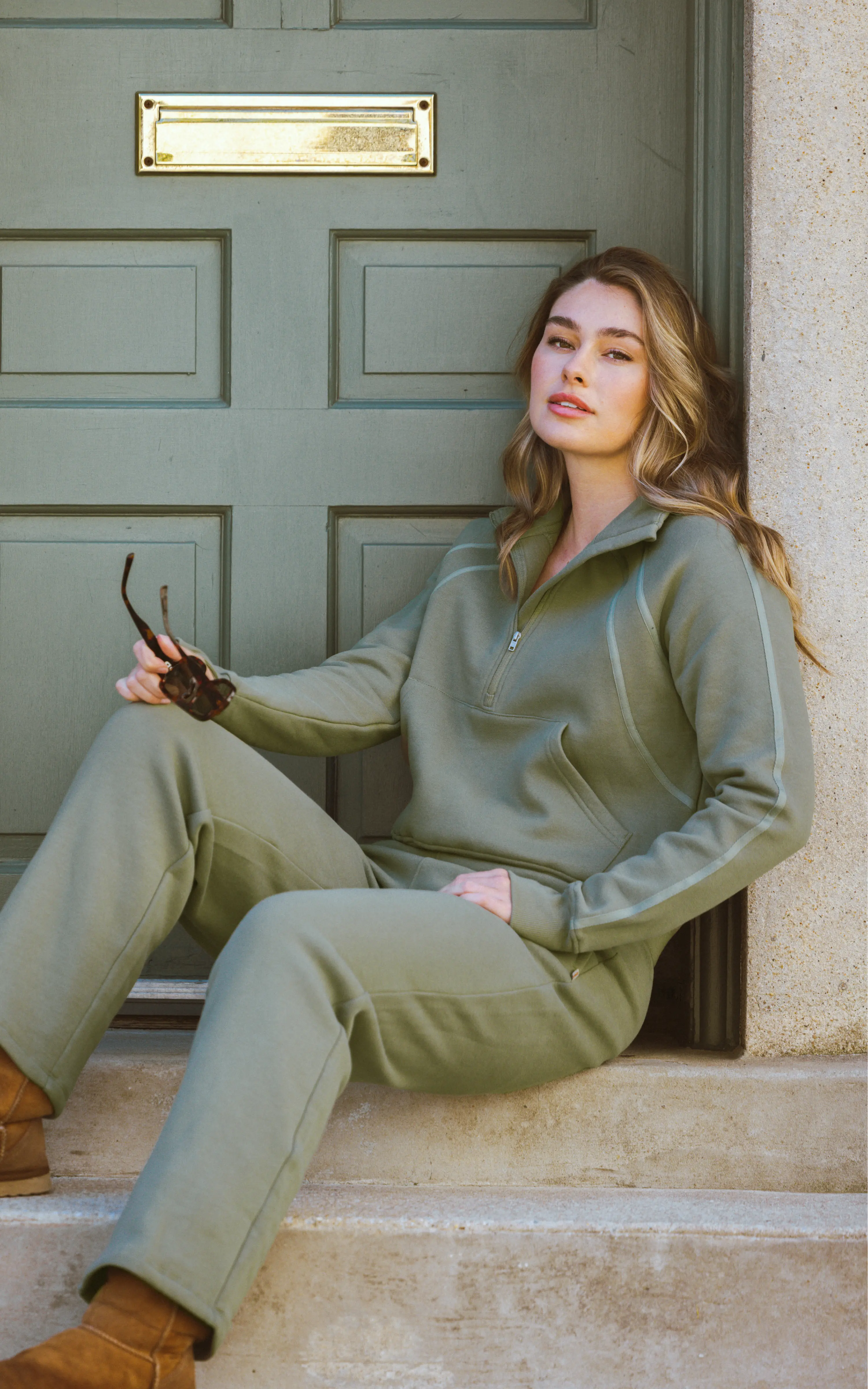 Olive green fleece half-zip pullover and straight-leg pants with a relaxed fit, modeled seated by a paneled door.