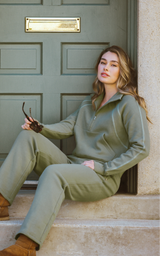 Olive green fleece half-zip pullover and straight-leg pants with a relaxed fit, modeled seated by a paneled door.
