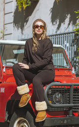 Woman wearing a dark brown plush crewneck and tapered pant set with cream shearling-lined cuffs and tan boots, seated on a.