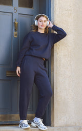 Woman wearing a navy fleece pullover and matching joggers with white sneakers, standing by a blue door.