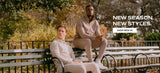 Two men in matching light beige casual lounge sets sitting on a dark green park bench with autumn foliage in the background. White text on the right reads 'NEW SEASON. NEW STYLES.' with a white 'SHOP NEW IN' button below it.