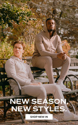 Two men in matching light beige casual lounge sets sitting on a dark green park bench with autumn foliage in the background. White text at the bottom reads 'NEW SEASON. NEW STYLES.' with a white 'SHOP NEW IN' button below it.